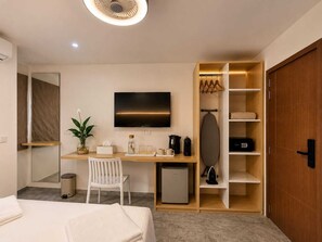 A hotel room with a bed, a desk with a computer, a TV, a small fridge, a plant, and a wardrobe.