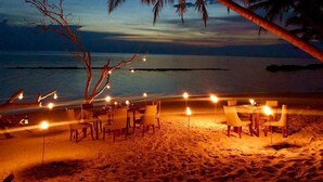 Montaje de comedor frente a la playa, con mesas y sillas bajo una palmera, con vista al océano.