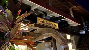 A wooden sign with "Veli Beach Inn" written on it, hanging under a wooden structure with a fan and lights.