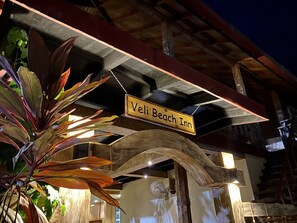 A wooden sign with "Veli Beach Inn" written on it, hanging under a wooden structure with a fan and lights.