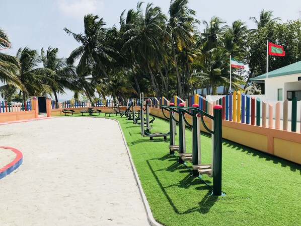 A playground with exercise equipment, a sandy area, and palm trees.