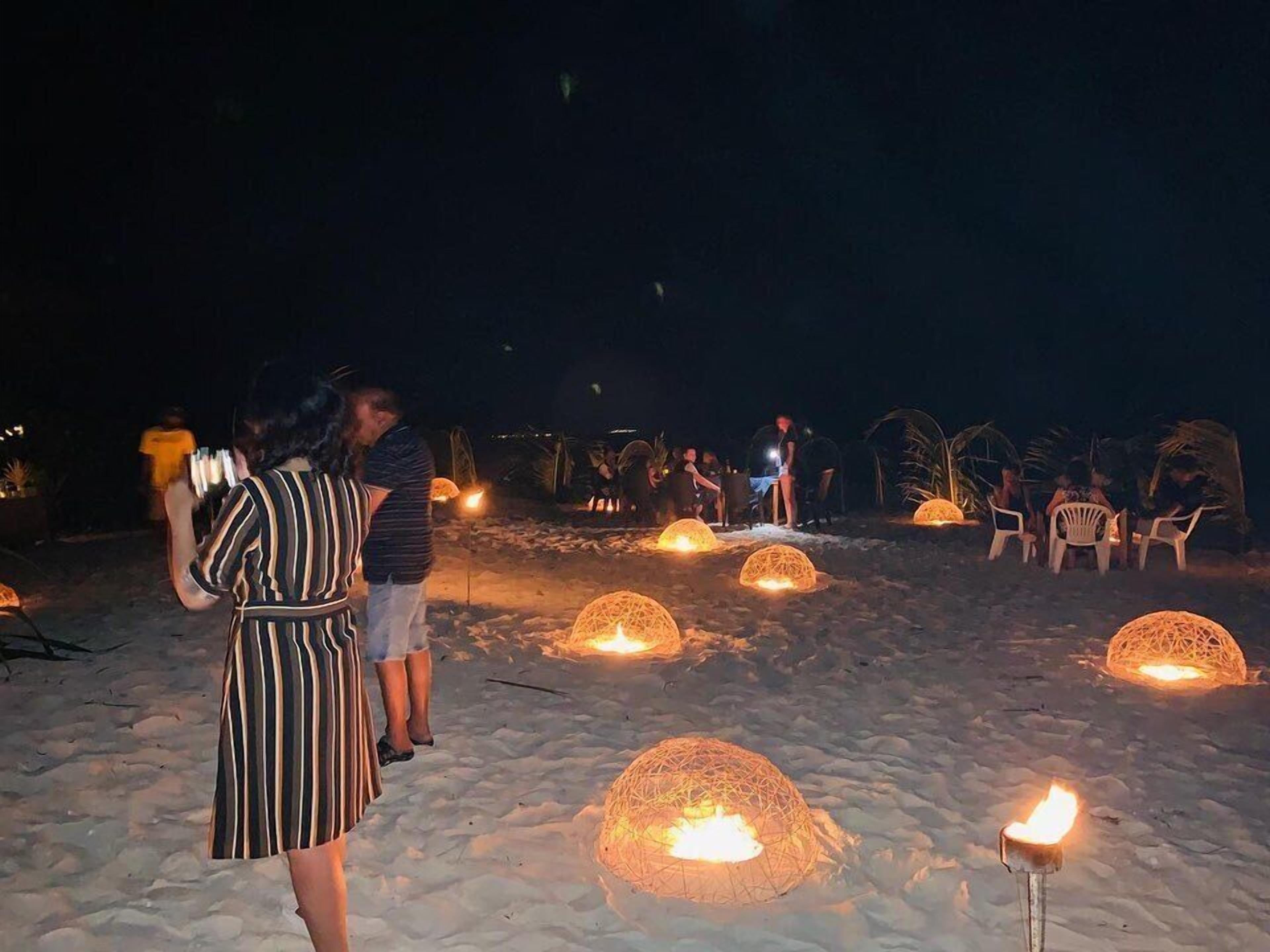 Escena de playa con gente reunida alrededor de estructuras esféricas con llamas, posiblemente para un evento social.