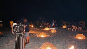 Escena de playa con gente reunida alrededor de estructuras esféricas con llamas, posiblemente para un evento social.