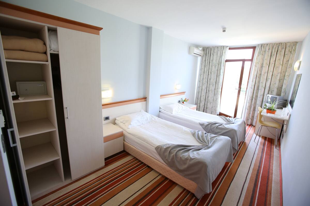 A hotel room with two beds, a wardrobe, a desk with a chair, and a window with curtains.