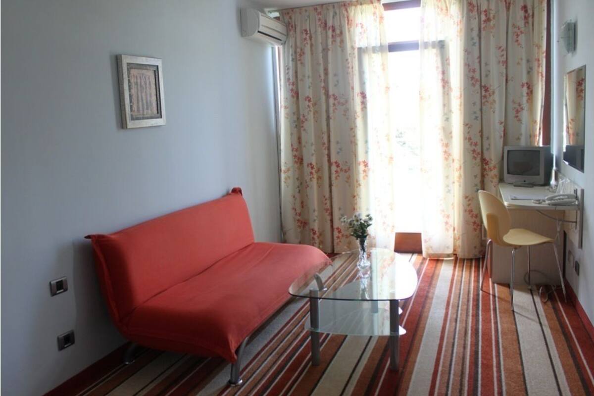 A room with a red sofa, a glass coffee table, a small desk with a chair, a television, and a window with floral curtains.