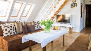 A loft-style living room with a wooden staircase, a sofa with patterned cushions, a dining table with a vase of flowers, and a television on a white table. - Mistral Dom Wakacyjny (Ustka)