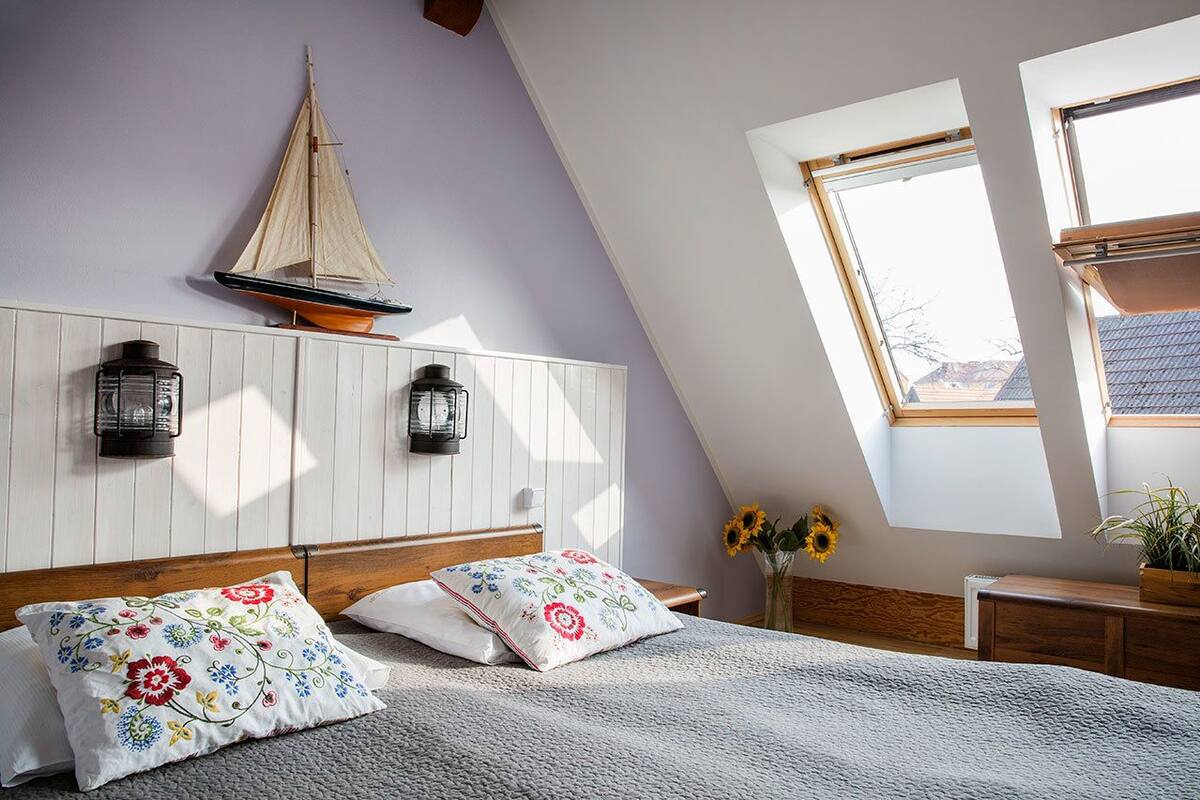 A bedroom with a sloped ceiling, a bed with floral pillows, a small sailboat decoration, two wall-mounted lanterns, and a window with a view of the outside.
