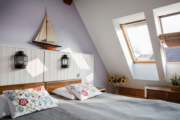A bedroom with a sloped ceiling, a bed with floral pillows, a small sailboat decoration, two wall-mounted lanterns, and a window with a view of the outside.