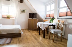 A bedroom with a bed, a TV on a wooden cabinet, a dining table with a chair, and a window with blinds.