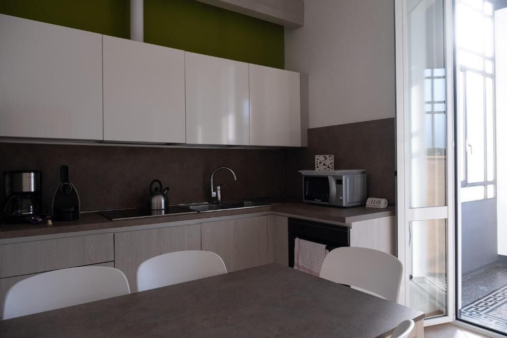 Une cuisine moderne avec des meubles blancs, un évier, une bouilloire et un four à micro-ondes. On y trouve une table à manger avec des chaises, et une porte vitrée offre une vue sur l’extérieur.