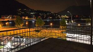 Ein nächtlicher Blick auf eine Uferpromenade mit Metallgeländer, einer Brücke im Hintergrund und beleuchteten Gebäuden an einem Hügelhang.