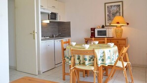 Une cuisine compacte avec des meubles blancs, un four à micro-ondes et un petit réfrigérateur. On y trouve une table à manger avec des chaises et une nappe, ainsi qu’une lampe sur une table de chevet.