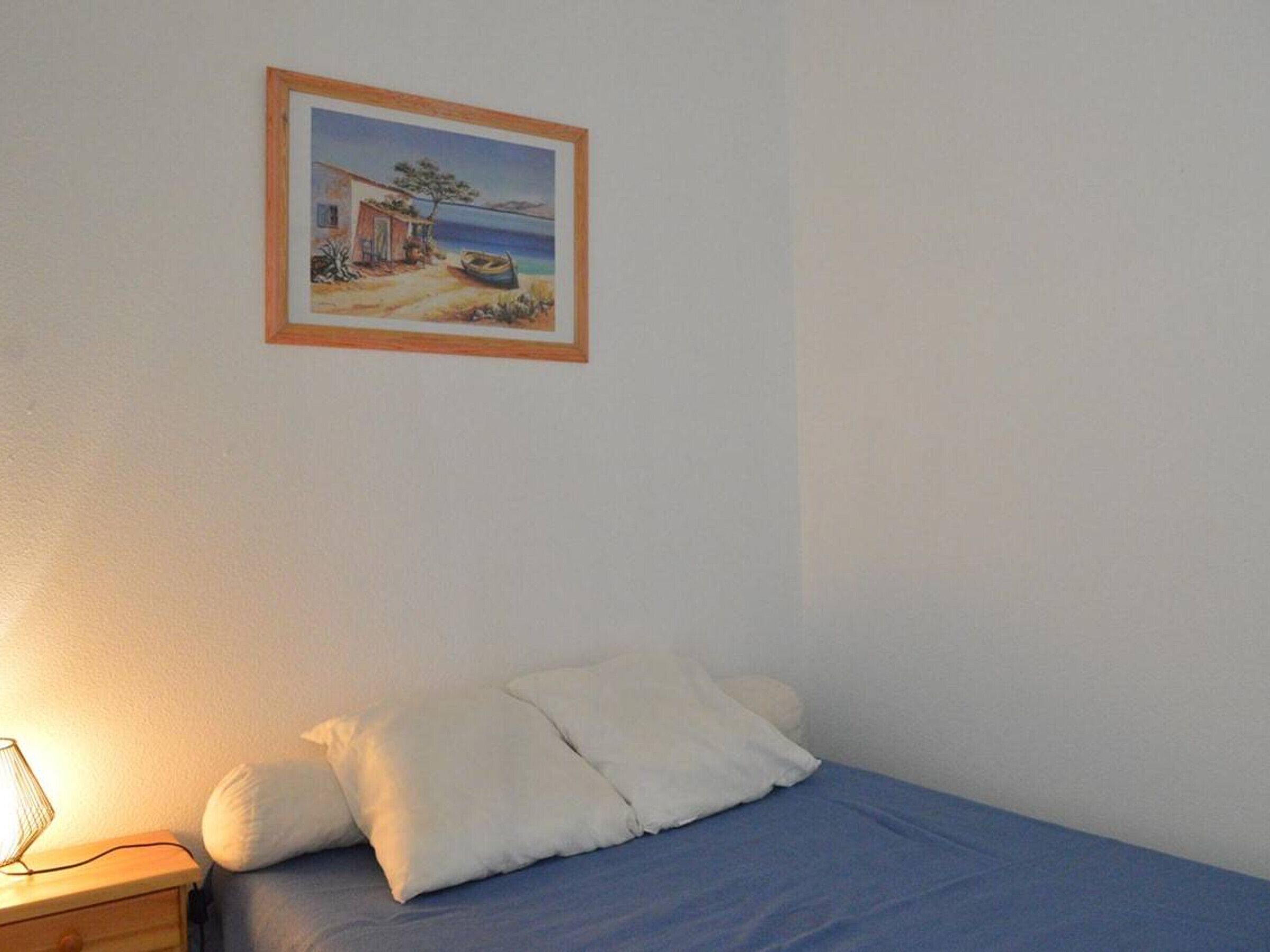 A bedroom with a bed, pillows, a bedside table with a lamp, and a framed picture on the wall.
