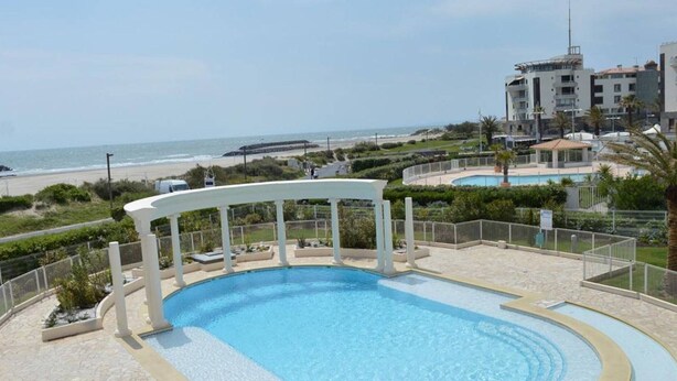 Une piscine agrémentée d’une pergola blanche, entourée d’une clôture, et offrant une vue sur la plage et l’océan en arrière-plan.