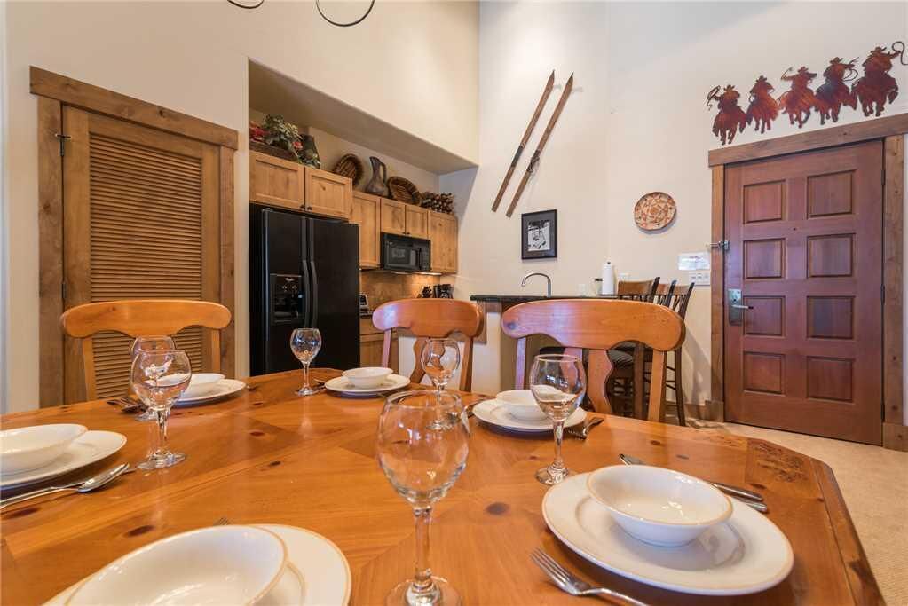 A dining area with a wooden table set for four, surrounded by wooden chairs. A kitchen with wooden cabinets and appliances is visible in the background.