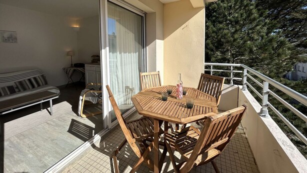 Een balkon met een houten tafel en stoelen, een schuifdeur en uitzicht op bomen.