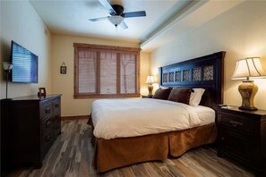 A bedroom with a bed, wooden floor, ceiling fan, window with blinds, bedside lamps, and a TV on a dresser. - 5111 Emerald Lodge, Trappeur's Crossing Apartment 2 (Steamboat Springs)