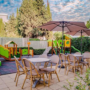 Un patio al aire libre con sillas de mimbre y una mesa, rodeado de juegos infantiles y una valla de madera.