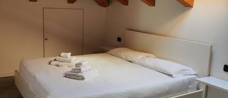A bedroom with a white bed, neatly made with towels, a white nightstand, and a wooden ceiling.