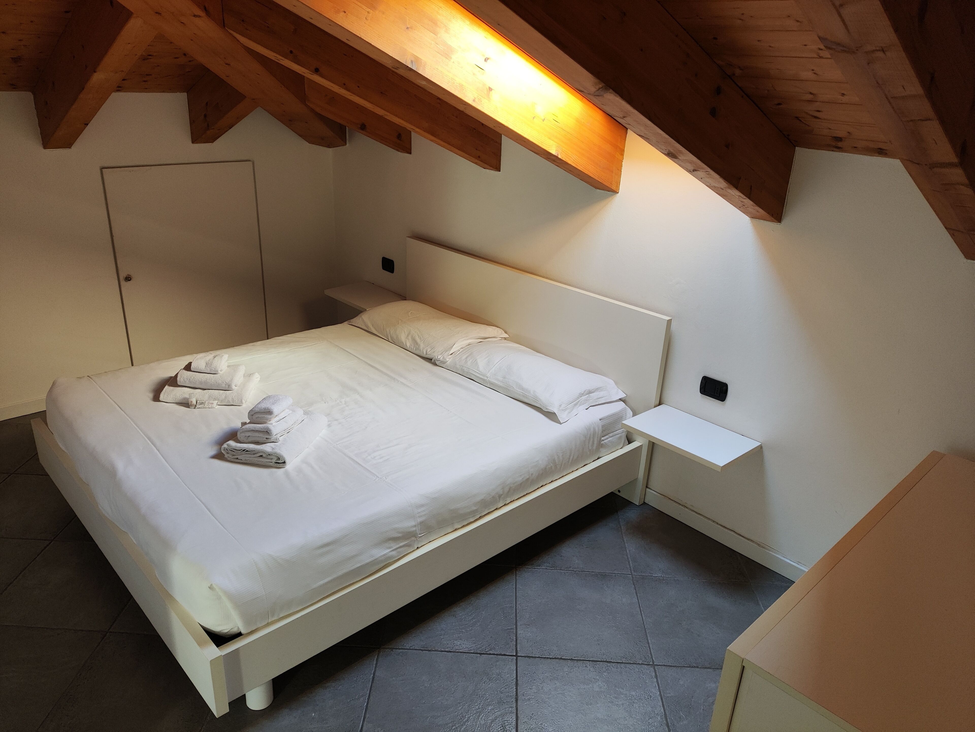 Quarto individual com uma cama, teto de madeira, paredes brancas e chão de azulejos.