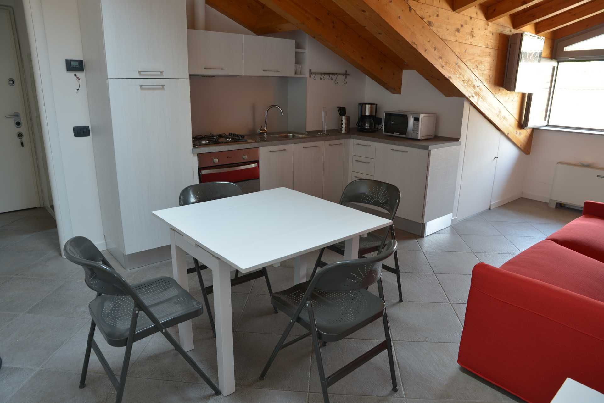 Et kompakt køkken med hvide skabe, en gasblus og en mikroovn. Der er et spisebord med fire stole og en rød sofa i opholdsstuen.