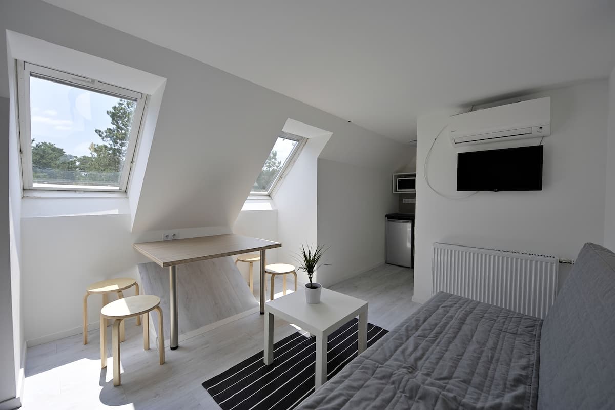 A modern, well-lit room with a loft-style ceiling, a bed, a small dining area, and a TV mounted on the wall.