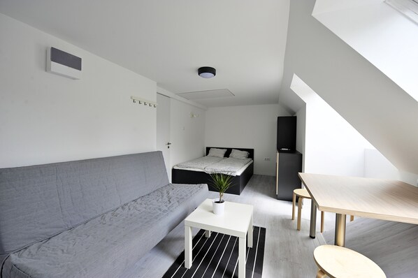 A modern, minimalist bedroom with a gray sofa, a small white table with a plant, a bed with a black headboard, and a wooden dining table.