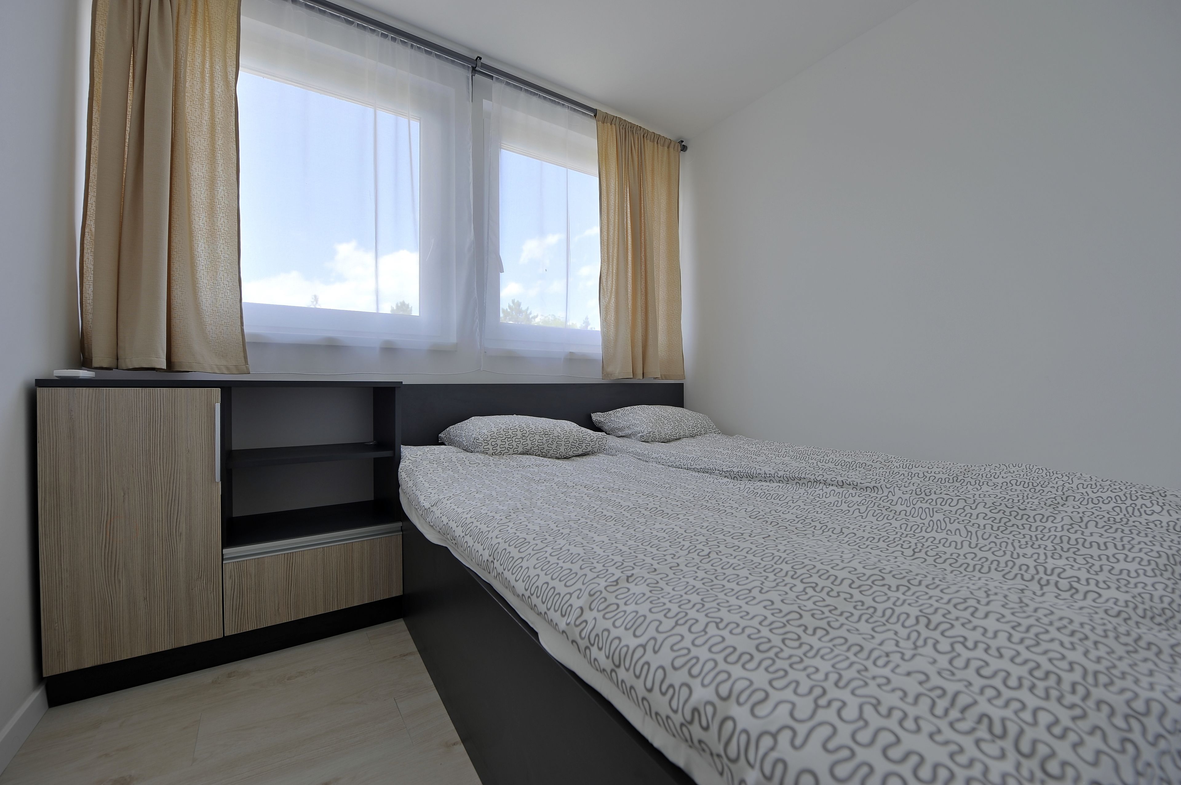 A bedroom with a bed, two windows with curtains, a built-in shelf, and a wooden floor.