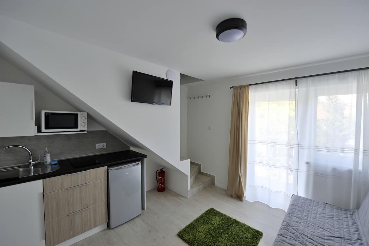 A modern kitchen with a microwave, a small refrigerator, and a sink. There is a staircase leading to a bedroom with a bed and a window with sheer curtains.