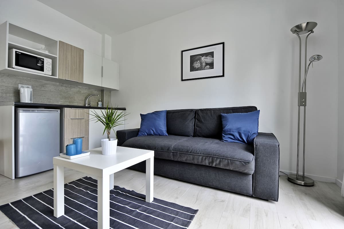 Ett modernt vardagsrum med en grå soffa, ett vitt bord, en svart- och vitrandig matta, en golvlampa och en köksdel med mikrovågsugn och kylskåp.
