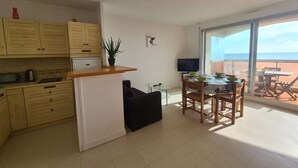 A modern kitchen with wooden cabinets, a dining area with a table and chairs, a TV, and a large sliding glass door leading to a balcony with a view of the beach. - Seafront 3-Room Apartment with Balcony, Pool, and Private Parking - Sleeps 4 (Saint-Jean-de-Monts)
