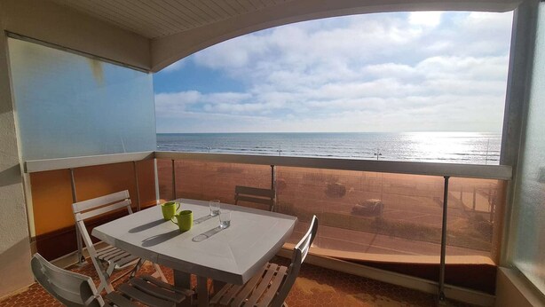 A balcony with a table and chairs, overlooking a beach with the sea and a cloudy sky. - Seafront 3-Room Apartment with Balcony, Pool, and Private Parking - Sleeps 4 (Saint-Jean-de-Monts)