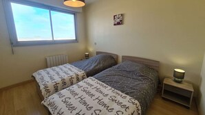A bedroom with two beds, a window, a radiator, a bedside table with a lamp, and a wall-mounted picture. - Seafront 3-Room Apartment with Balcony, Pool, and Private Parking - Sleeps 4 (Saint-Jean-de-Monts)