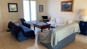 A cozy living room with a sofa, a chair, a coffee table, and a TV. There are two windows with curtains, a door, and a painting on the wall. - House near pond, enclosed garden, 2 bedrooms (Bretignolles-sur-Mer)