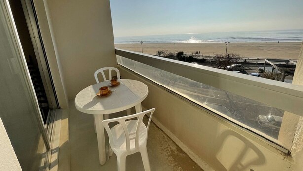 Un balcon avec une table et des chaises blanches, donnant sur une plage, avec un ciel dégagé et un soleil radieux.