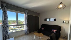 A modern living room with a black sofa, a small wooden table, and a view of a cityscape through large windows. - Beautiful apartment with parking near the beach, 2 rooms, 4 people (Saint-Jean-de-Monts)