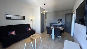A compact living space with a black sofa, a small wooden table, a dining area with chairs, and a kitchenette with a microwave. - Beautiful apartment with parking near the beach, 2 rooms, 4 people (Saint-Jean-de-Monts)