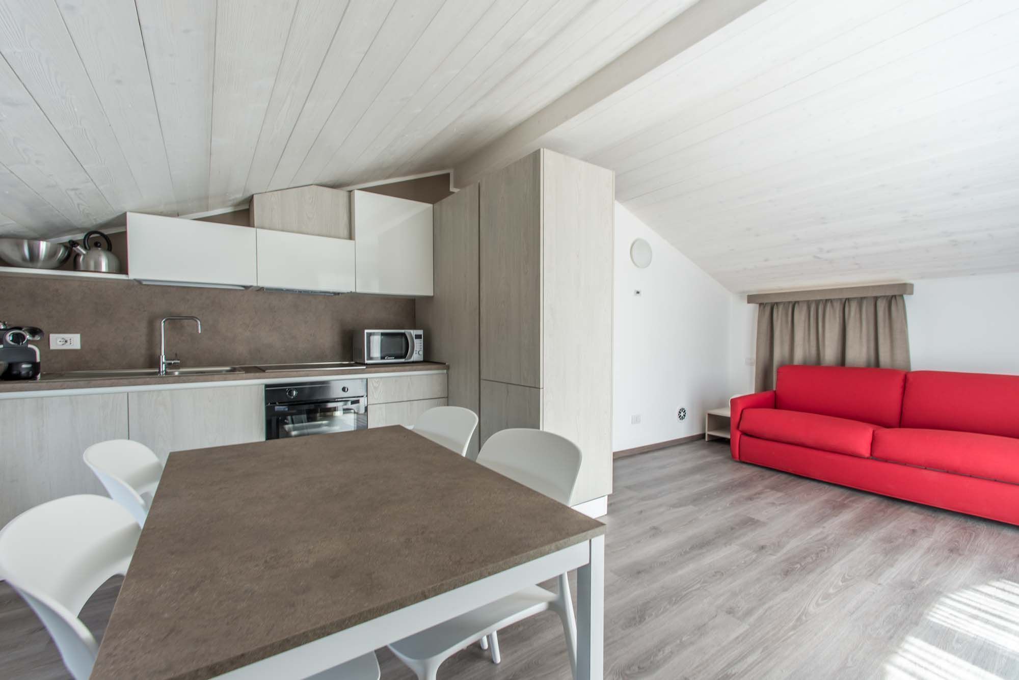 A modern kitchen with white cabinets, a microwave, and a sink. There is a dining table with chairs and a red sofa in the living area.