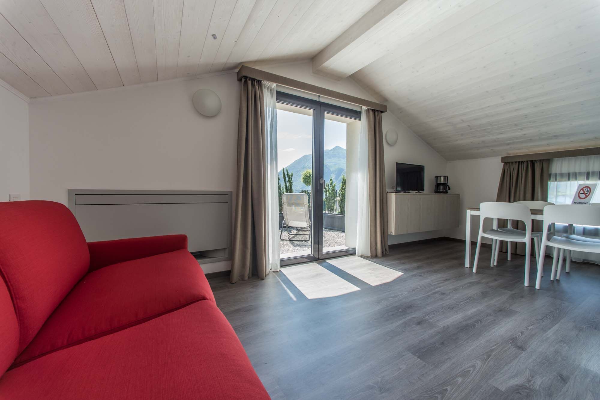 A modern, well-lit room with a red sofa, a dining area with a table and chairs, a TV, and a sliding glass door leading to an outdoor area with a mountain view.