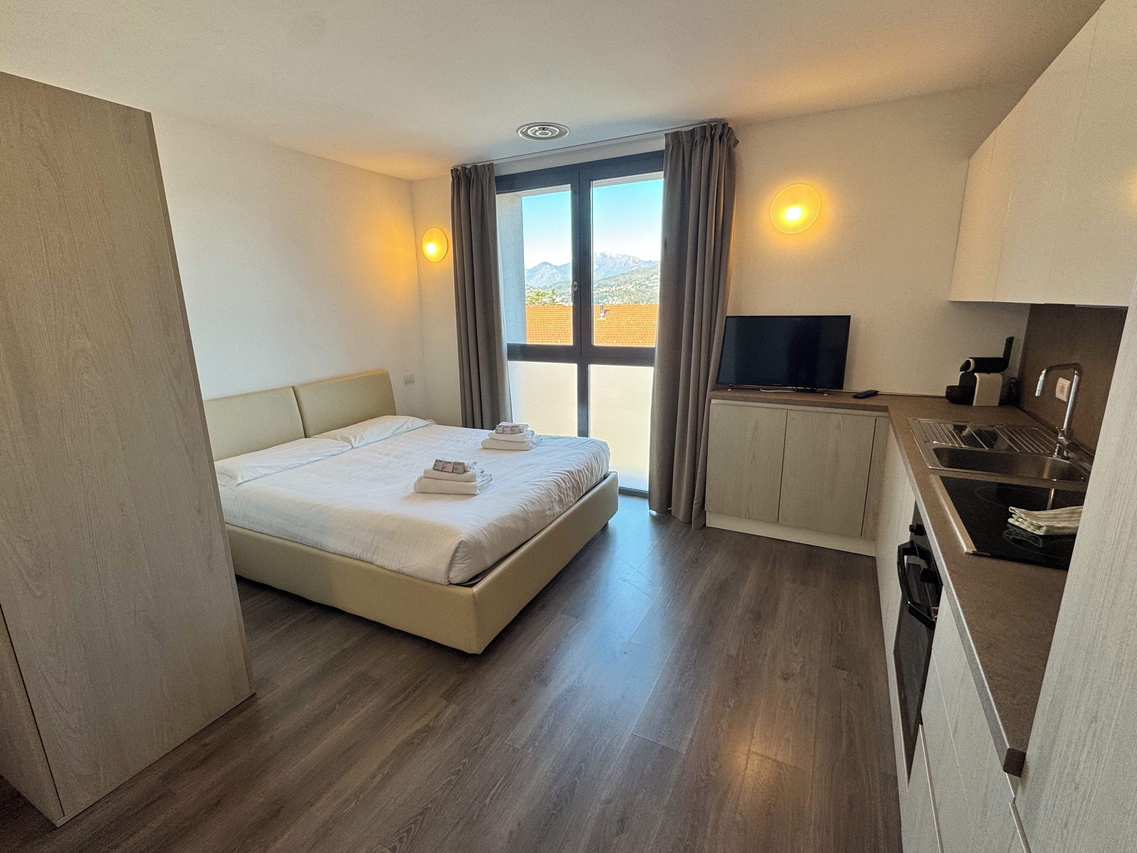 A hotel room with a bed, a TV, a kitchenette, and a view of the mountains.