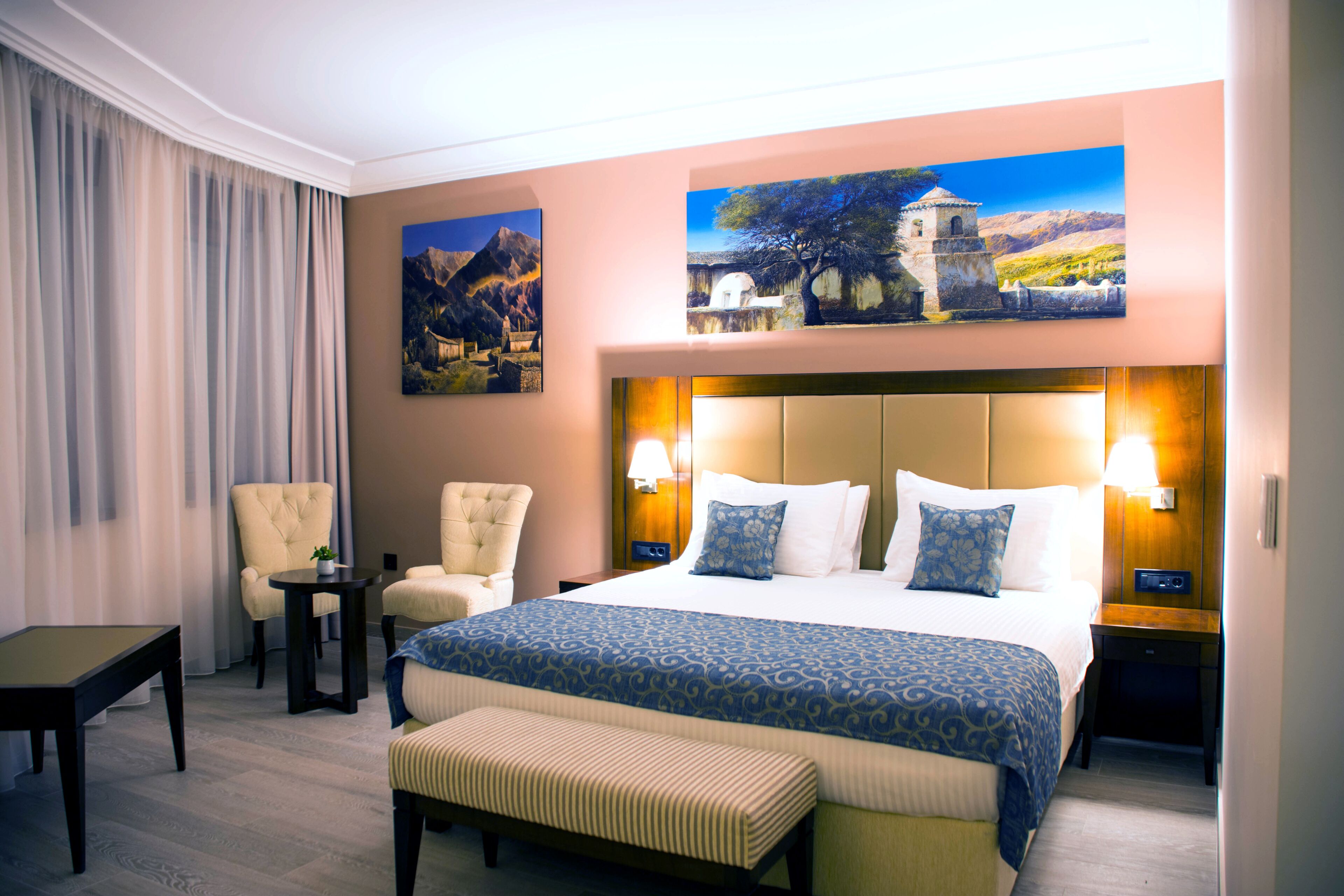 A hotel room with a large bed, two armchairs, a small table, and a bench. There are two paintings on the wall and a lamp on the nightstand.