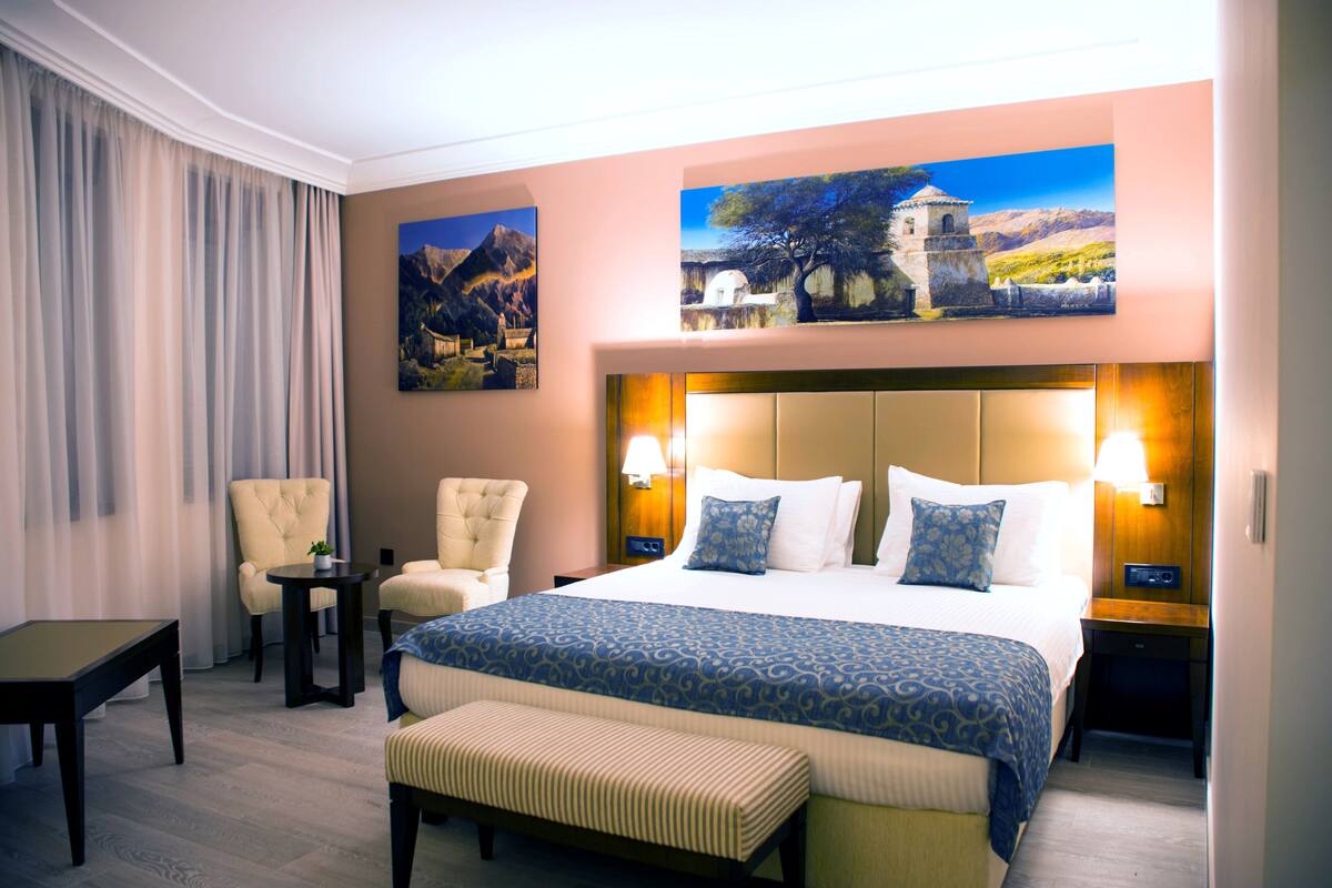 Habitación de hotel con una cama grande, dos sillones, una mesita y un banco. Hay dos lámparas de pared y dos cuadros sobre la cama.