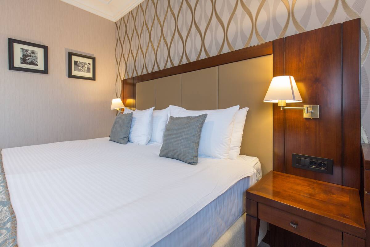 Una cama bien hecha con ropa de cama blanca y almohadas grises, una mesita de noche de madera con una lámpara, y dos cuadros en la pared.