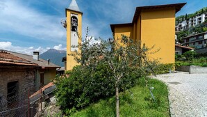 A yellow building with a bell tower and a cross, surrounded by greenery and a mountain in the background. - Regolo House (Perledo)