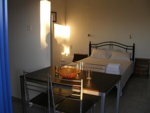 A hotel room with a bed, a bedside table, a table with chairs, and a lamp. - Panoramakamil (Faistos)