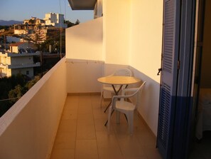 A balcony with a small table and chairs, a door, and a view of buildings in the background. - Panoramakamil (Faistos)