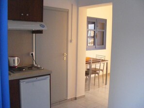 Eine kompakte Küche mit einem Kühlschrank, einem Wasserkocher und einer Mikrowelle. Durch eine offene Tür ist ein Essbereich mit Tisch und Stühlen zu sehen.