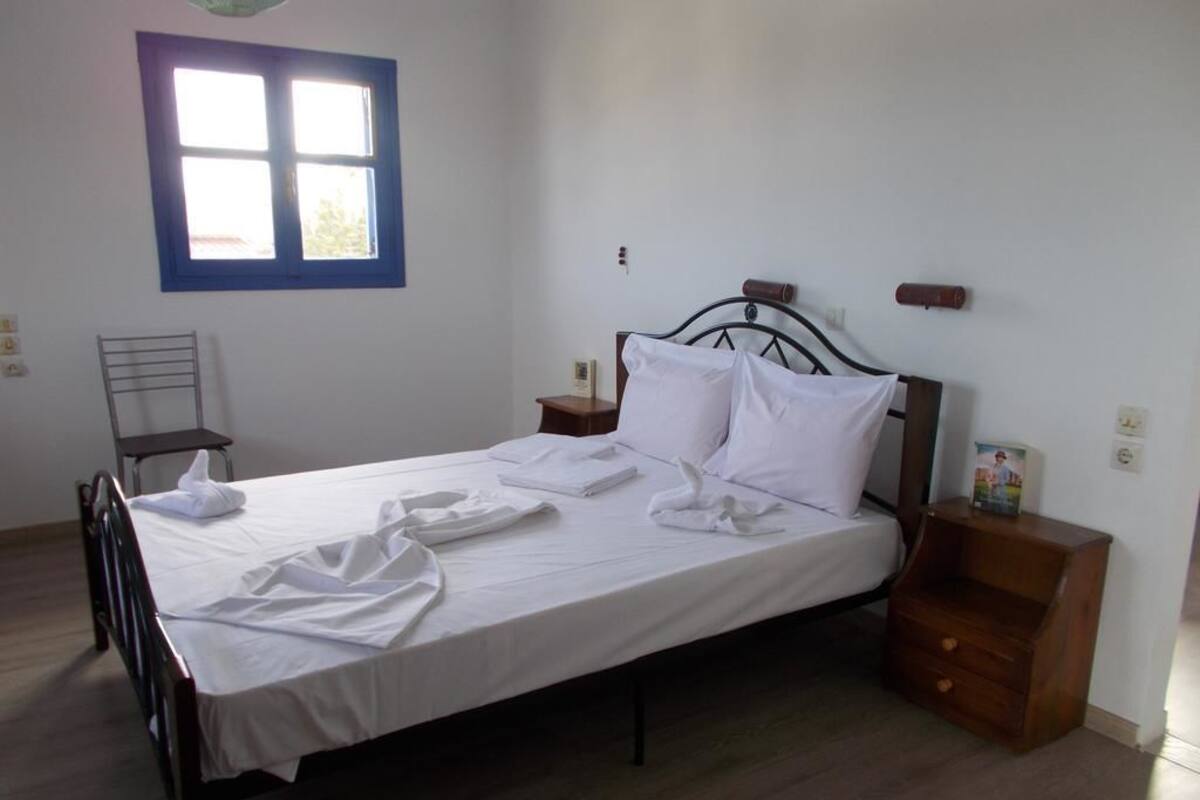 A single bed with a metal frame, a wooden nightstand, a chair, and a window with a blue frame.