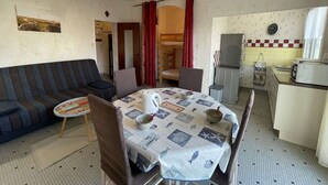 Ett kompakt bostadsutrymme med en soffa, ett matbord med en mönstrad duk, en köksdel med kylskåp och mikrovågsugn samt ett litet fönster.