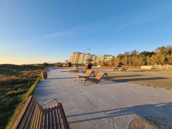 A paved outdoor area with multiple lounge chairs and a wooden bench, surrounded by a low wall and vegetation.